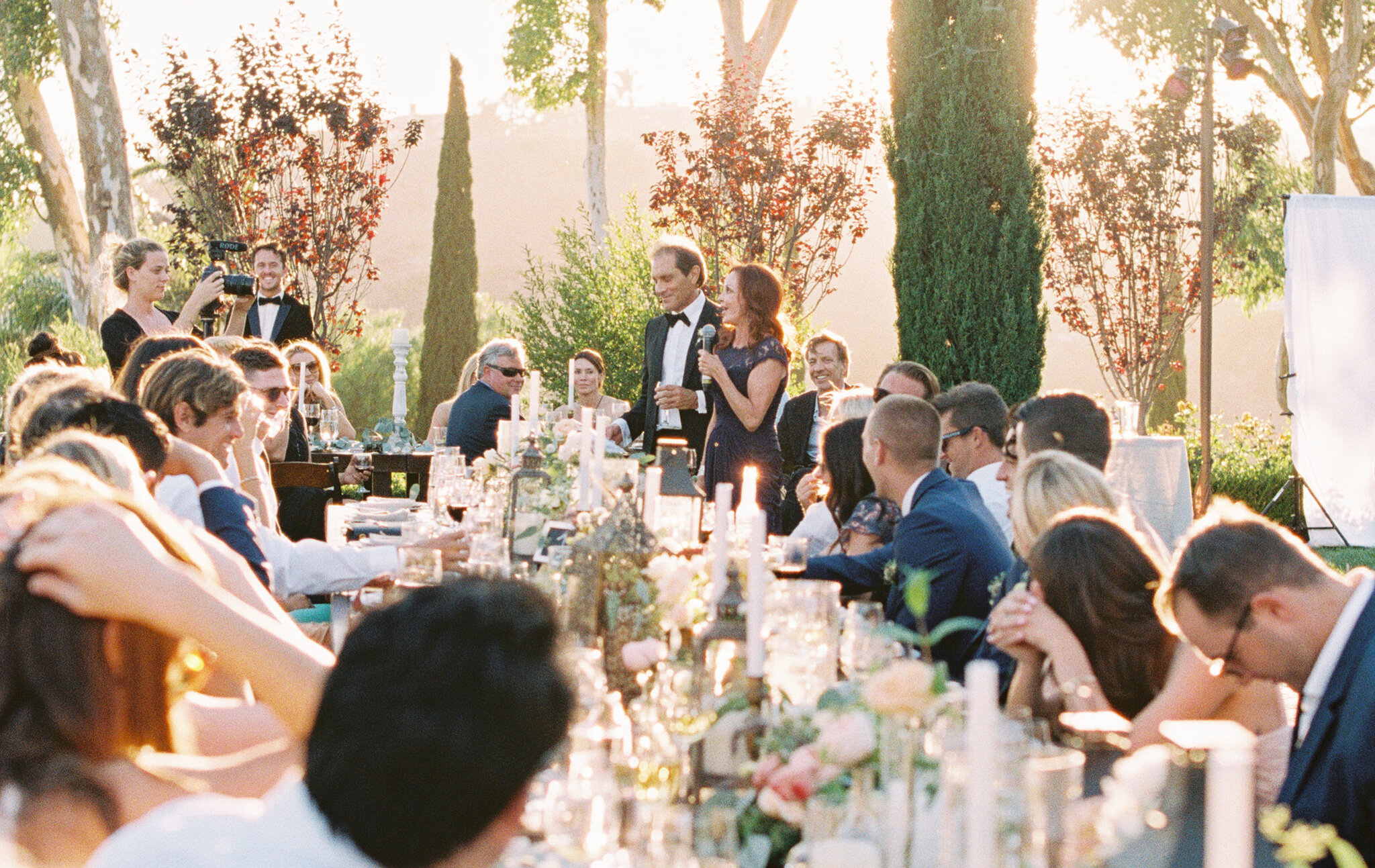 Guests at an outdoor wedding reception sitting at long tables decorated with candles and flowers as a couple gives a speech. The setting is lush with trees and greenery, with the sun setting in the background.