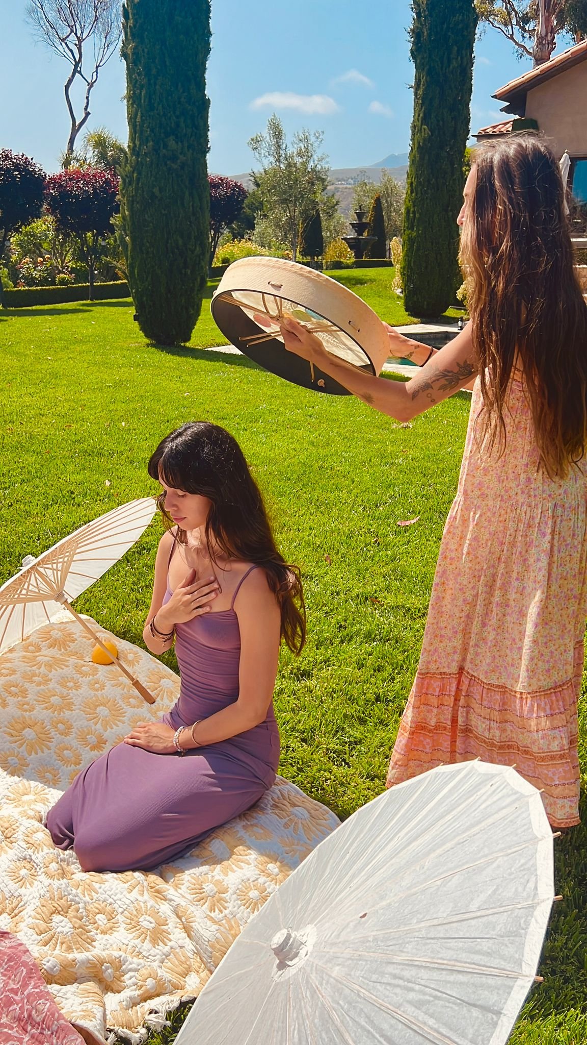 A woman kneeling on a blanket with white and yellow daisy pattern, holding her chest, as another woman stands holding a large umbrella over her head on a sunny day in a lush garden. | www.mayaarmonyart.com