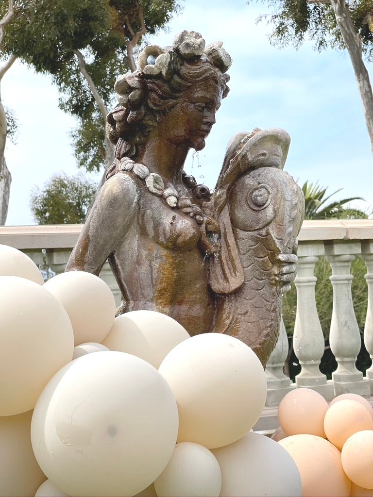 Stone sculpture of a woman holding a fish surrounded by white and peach balloons.