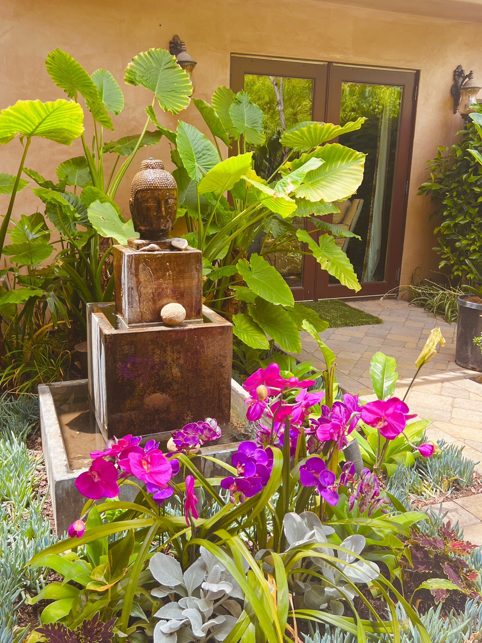 A garden with lush green plants, pink and purple flowers, and a small water fountain with a Buddha head sculpture, set against a background of a patio, a sliding glass door, and beige walls.