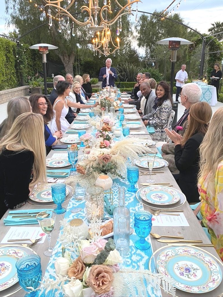 A large outdoor dining table set for a celebration with floral arrangements, blue glassware, gold utensils, and decorative plates, under a chandelier and string lights, with people dressed in semi-formal attire enjoying the event.