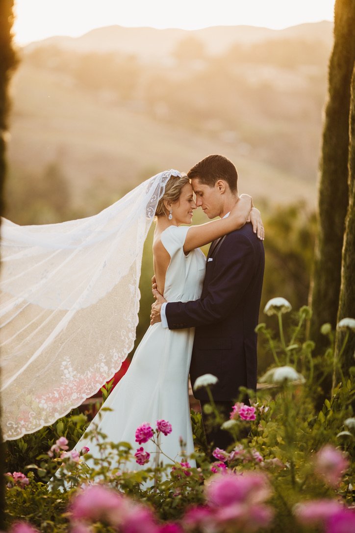 A bride and groom embrace in a lush garden at sunset, surrounded by pink and white flowers, with a scenic hilly landscape in the background.