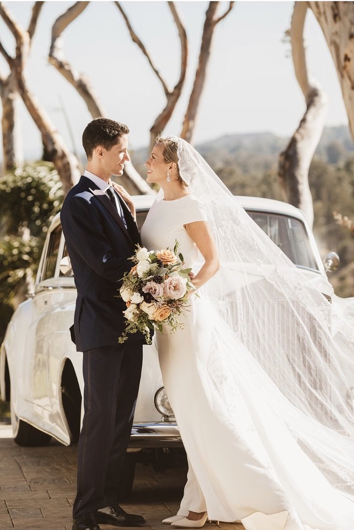 Bride and groom face to face in wedding attire outdoors with trees and hills in background, standing in front of a vintage car, the bride holding a large bouquet of flowers.