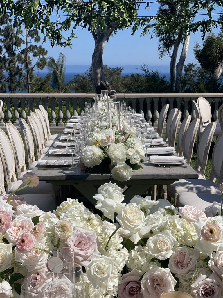 Outdoor dining setup with a long table decorated with white and blush flowers, surrounded by white chairs, overlooking trees and a distant body of water under a blue sky.