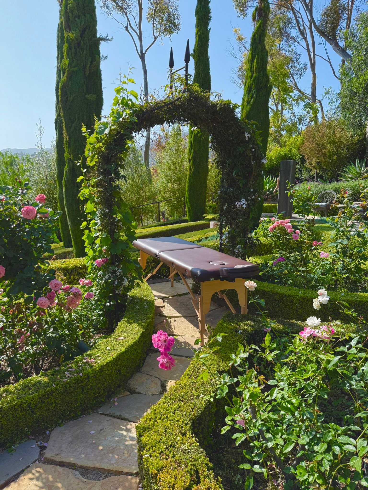 A massage table set up in a lush, vibrant garden with a stone pathway, an arched floral trellis, and tall green trees in the background.