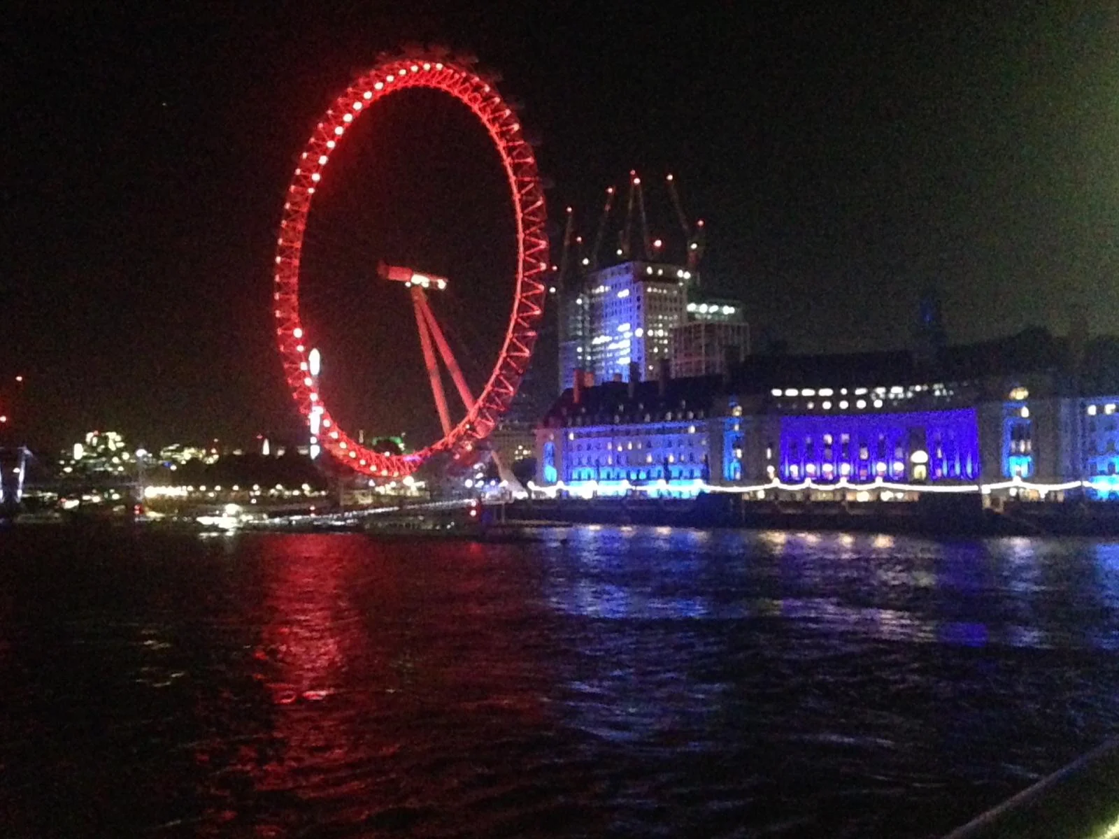 Night view of the London Eye illuminated in red, with surrounding buildings lit in blue, reflecting on the River Thames.