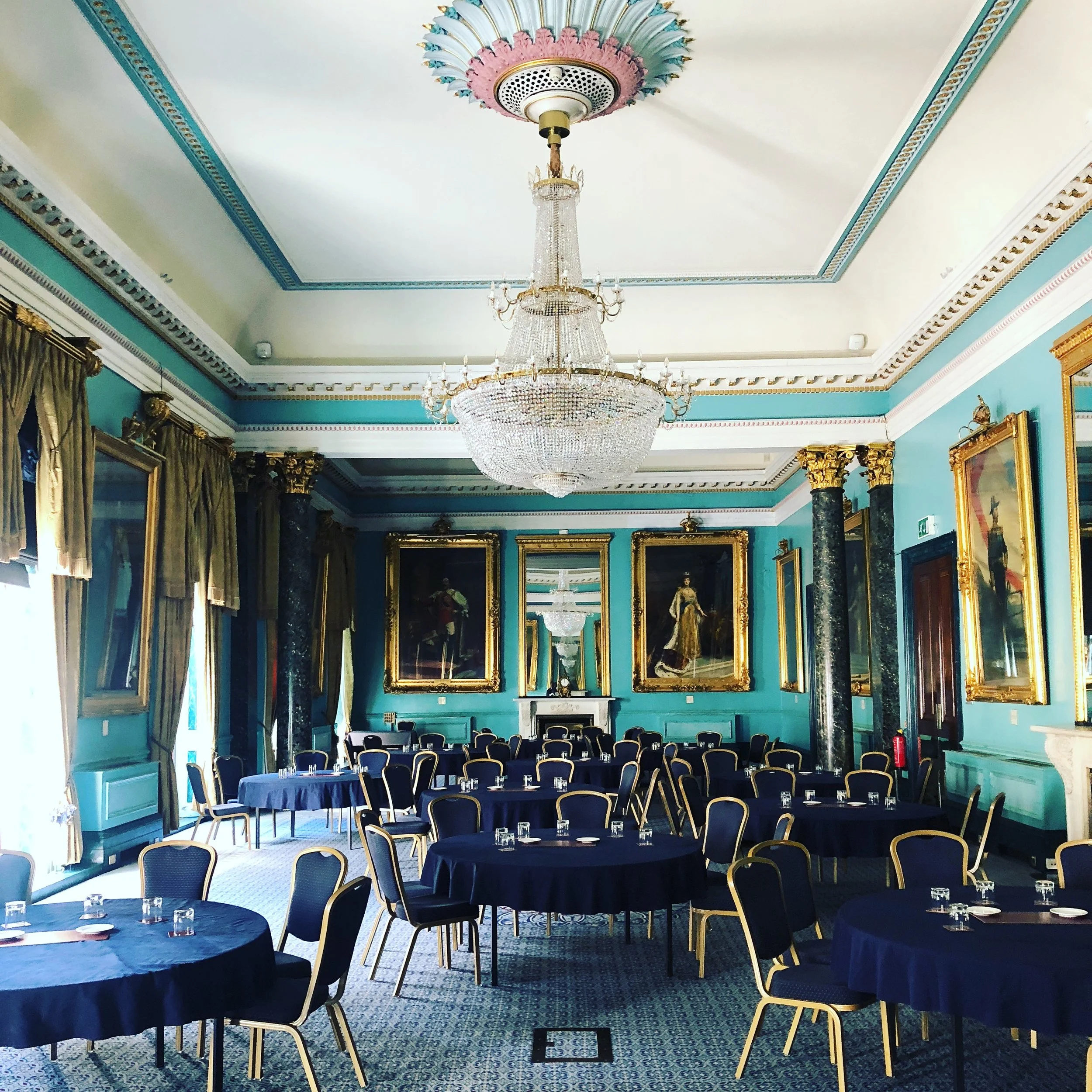 Elegant dining room with blue walls, round tables, navy tablecloths, ornate chandelier, and large framed portraits.