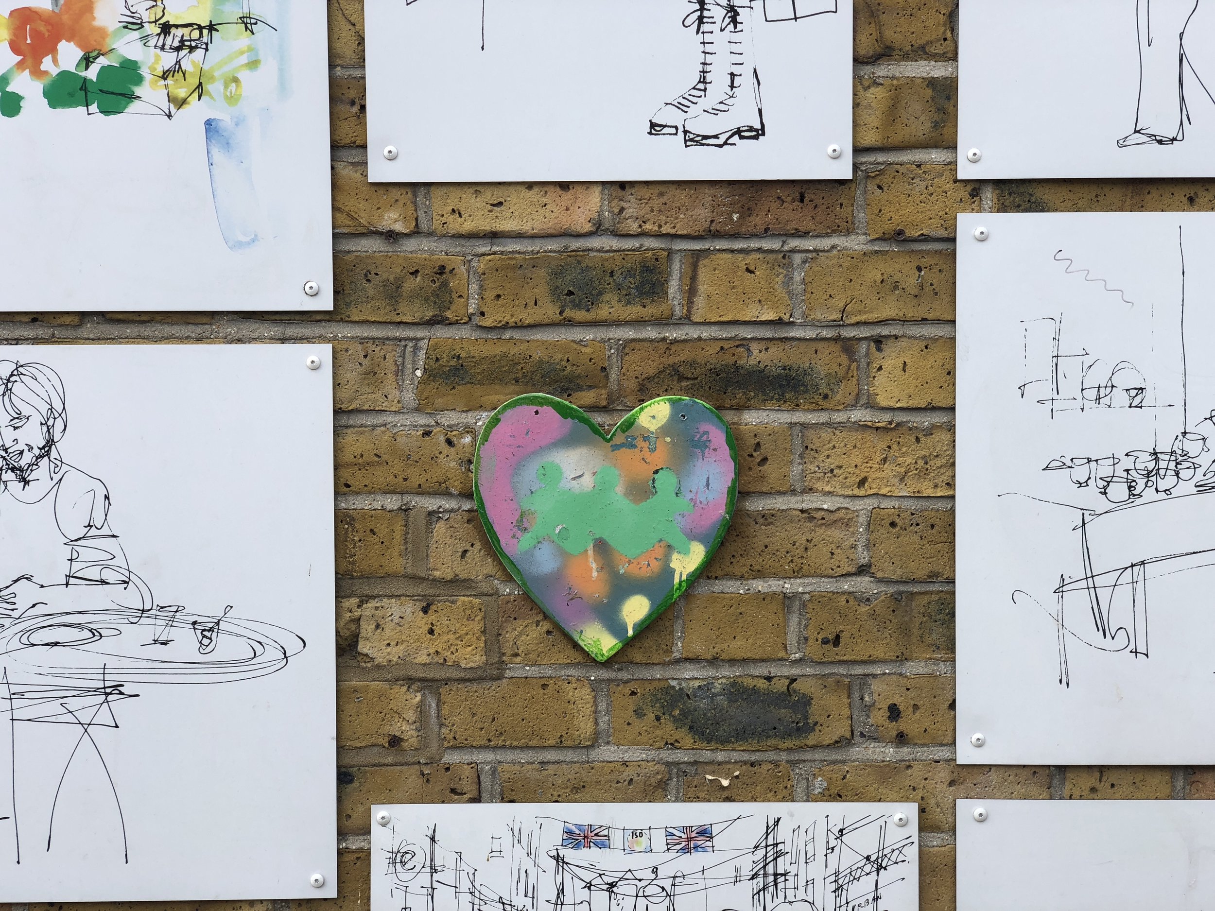 A colorful heart-shaped artwork displayed on a brick wall, surrounded by several white boards featuring abstract black line drawings of people and objects.