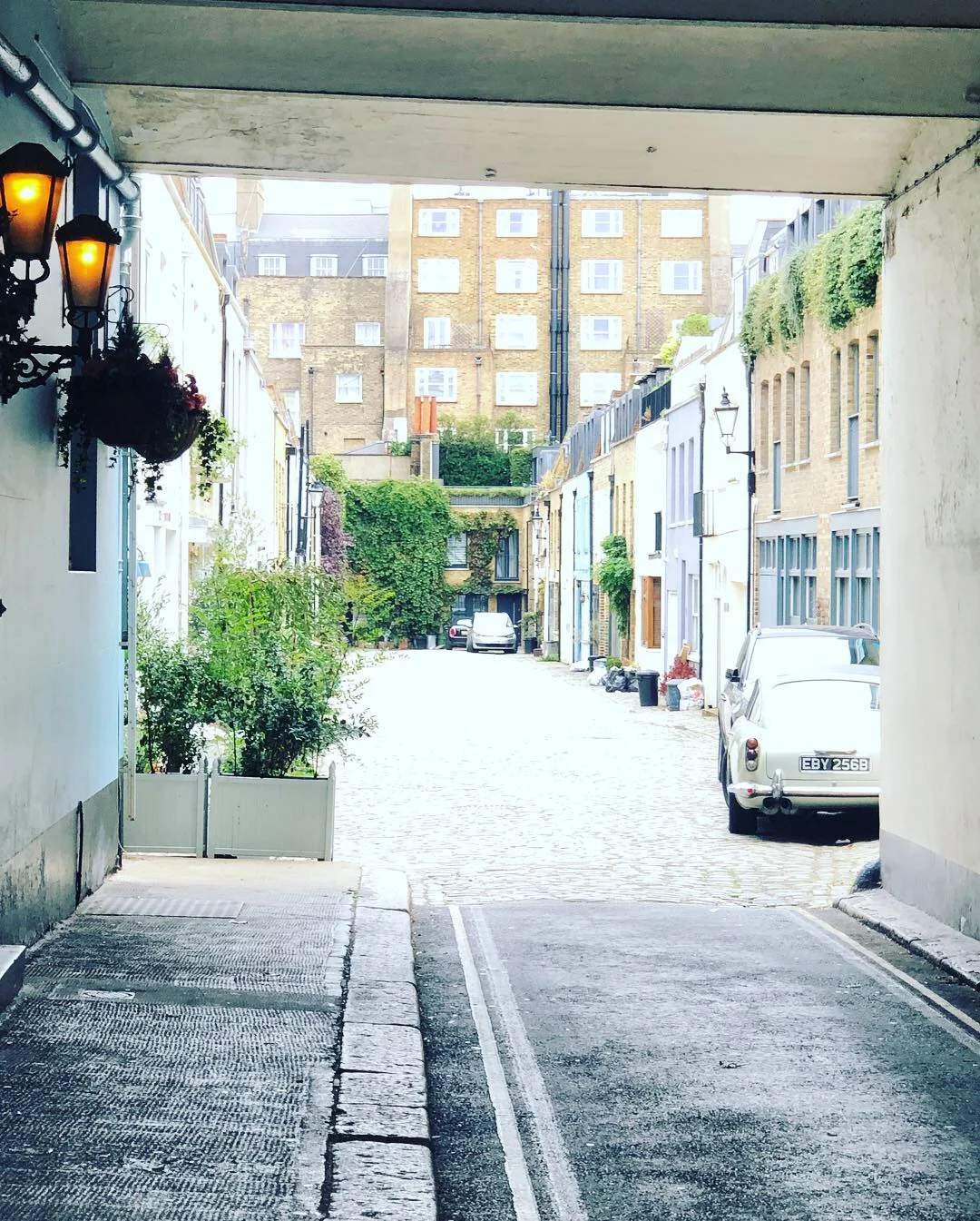 Cobbled street with vintage car, lined with brick buildings, plants, and hanging lanterns.