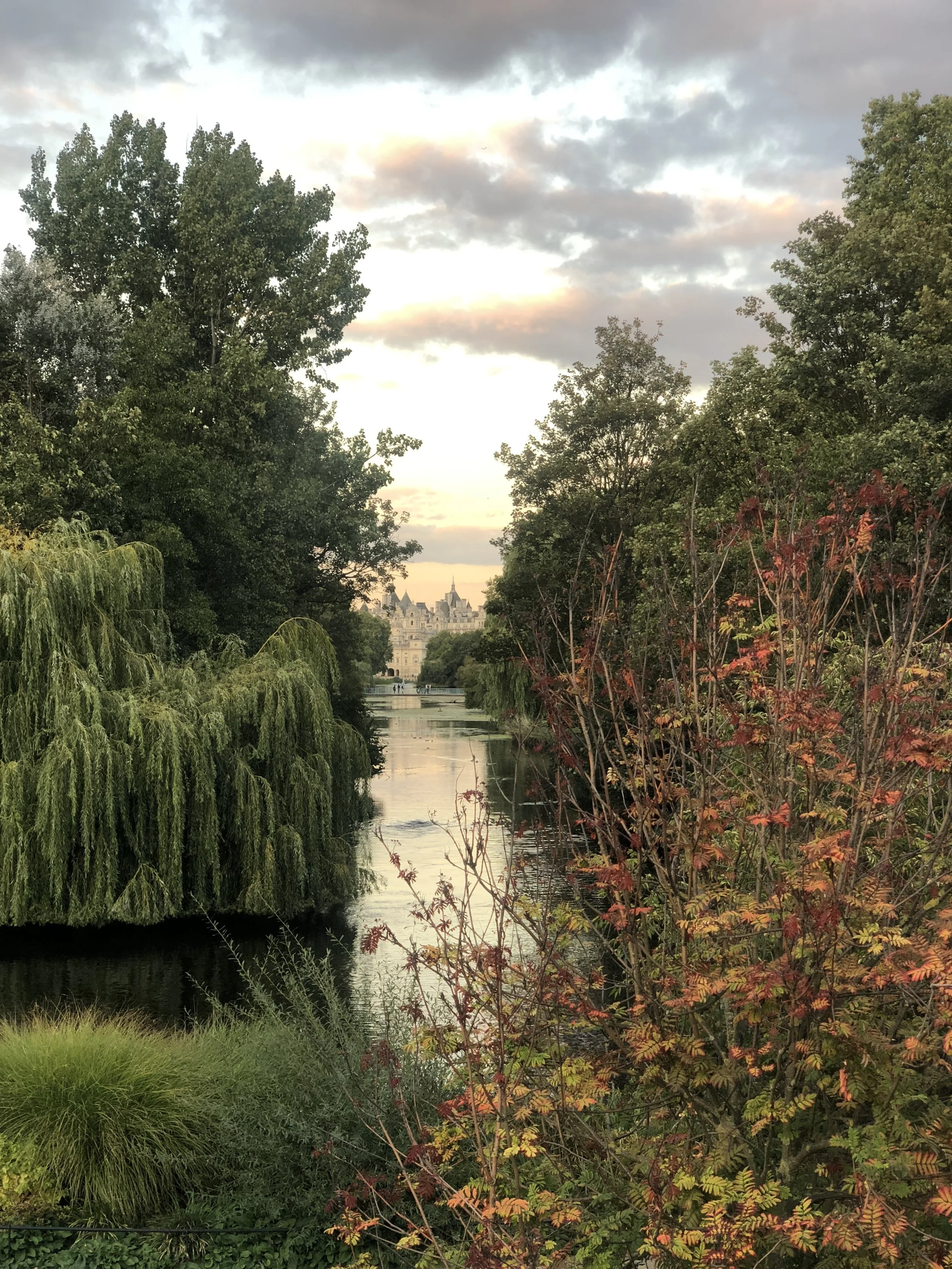 Scenic view of a tranquil lake surrounded by lush green trees and foliage, under a cloudy sky. In the distance, a historic building is visible across the water.