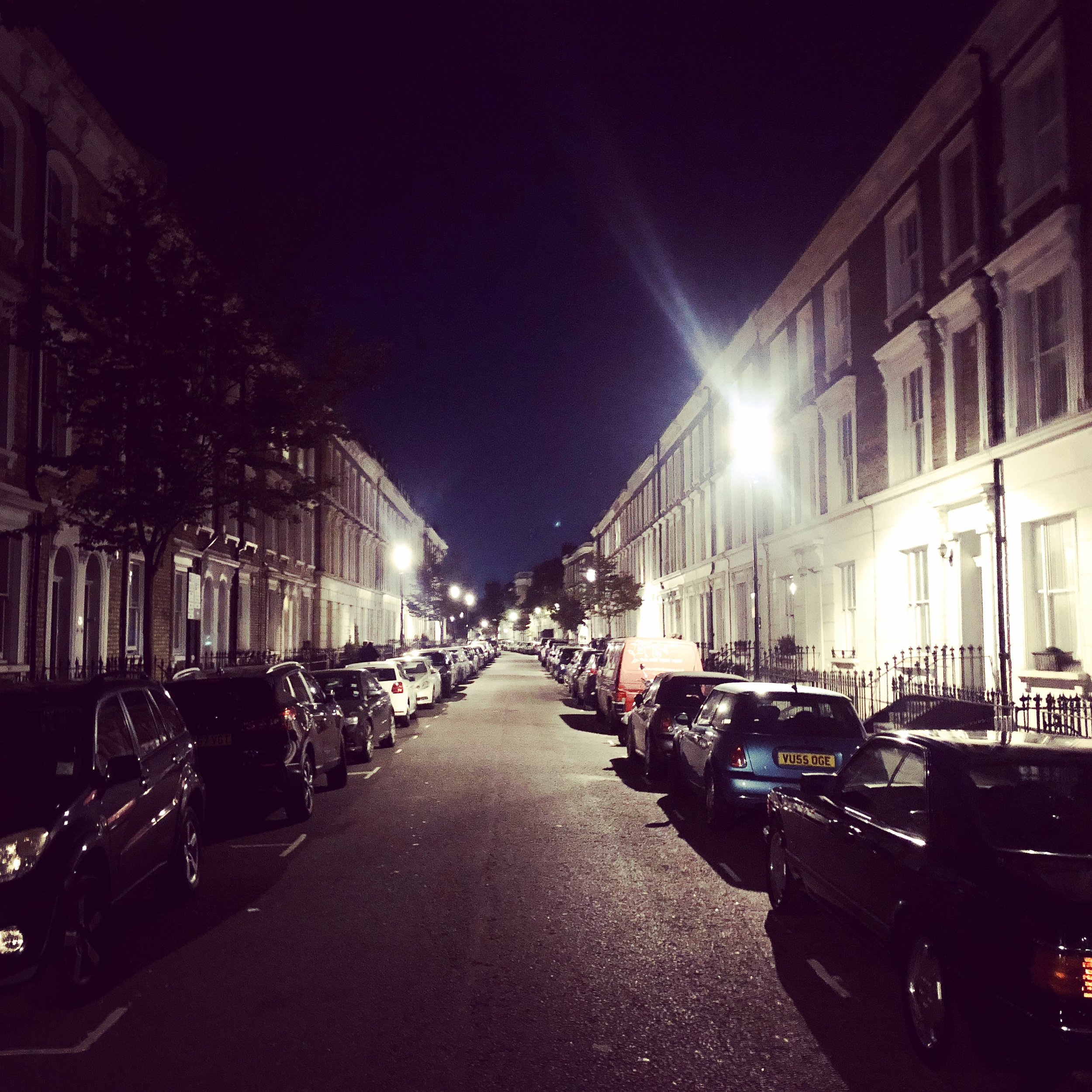 Night view of a quiet residential street lined with parked cars and illuminated by streetlights.