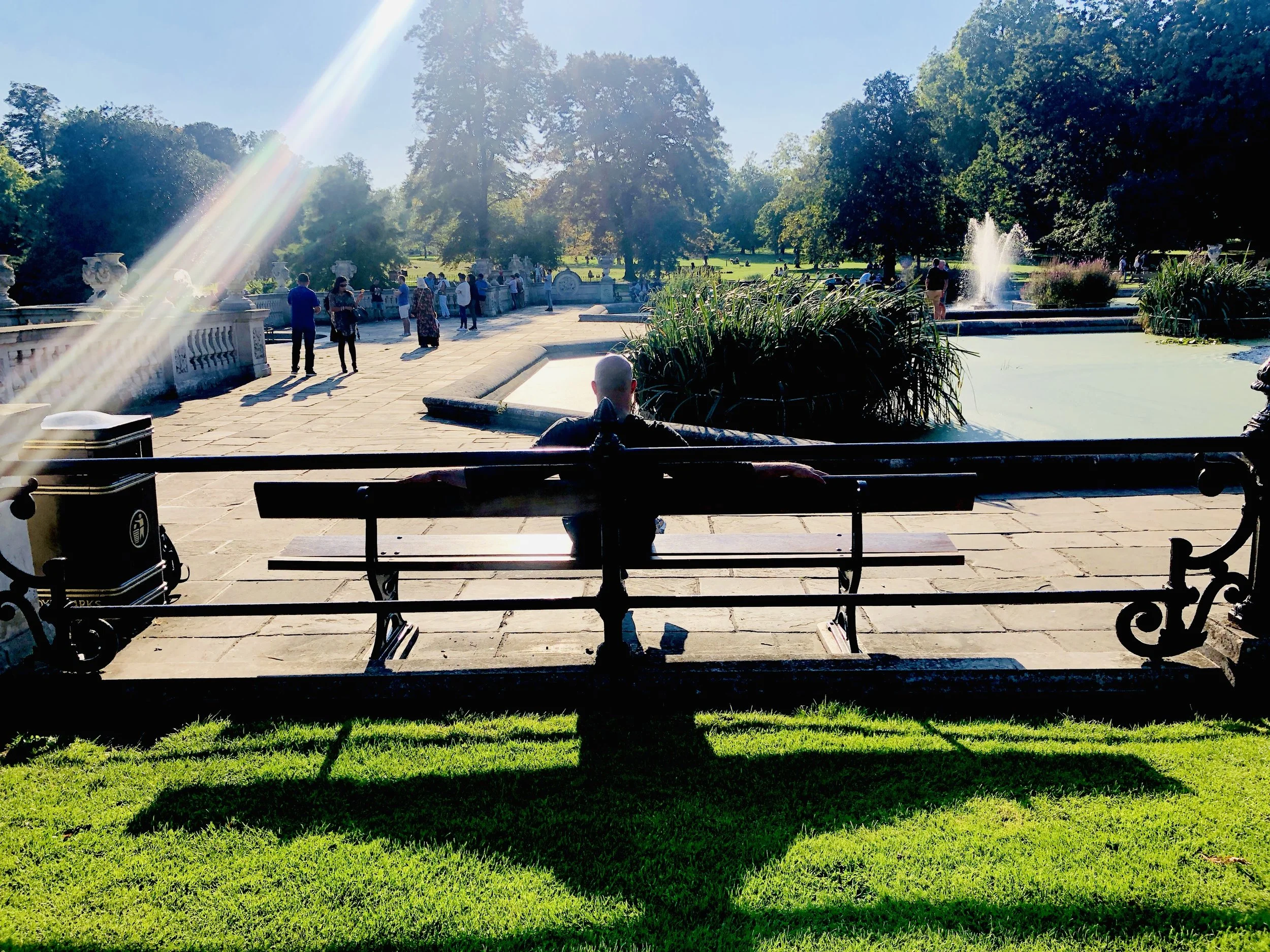 Person sitting on a bench in a park with a fountain and trees in the background, sunlight and shadows adding contrast to the scene.