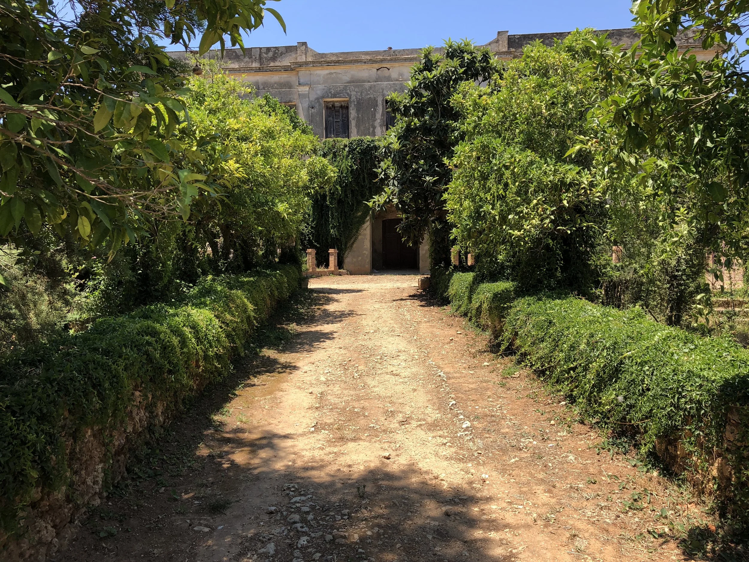 Pathway leading to an old stone building surrounded by green trees and bushes under a clear blue sky.