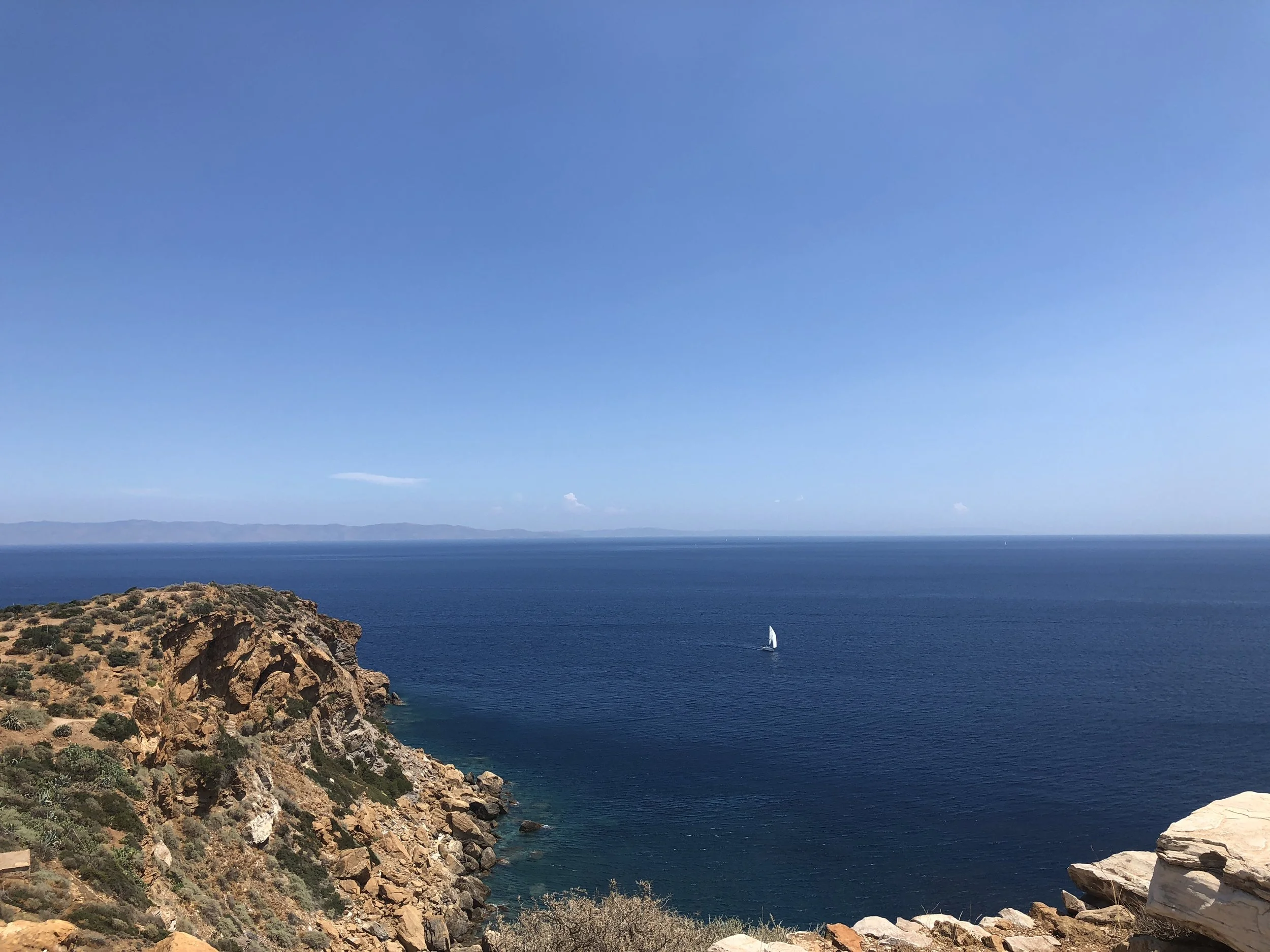 Rocky cliff overlooking a calm blue sea with a single white sailboat in the distance under a clear blue sky.
