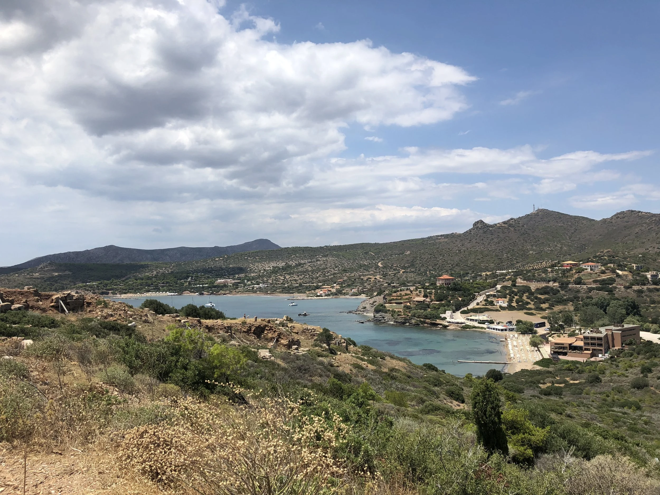 Coastal landscape with hills, bay, and beach in a seaside village under a partly cloudy sky.