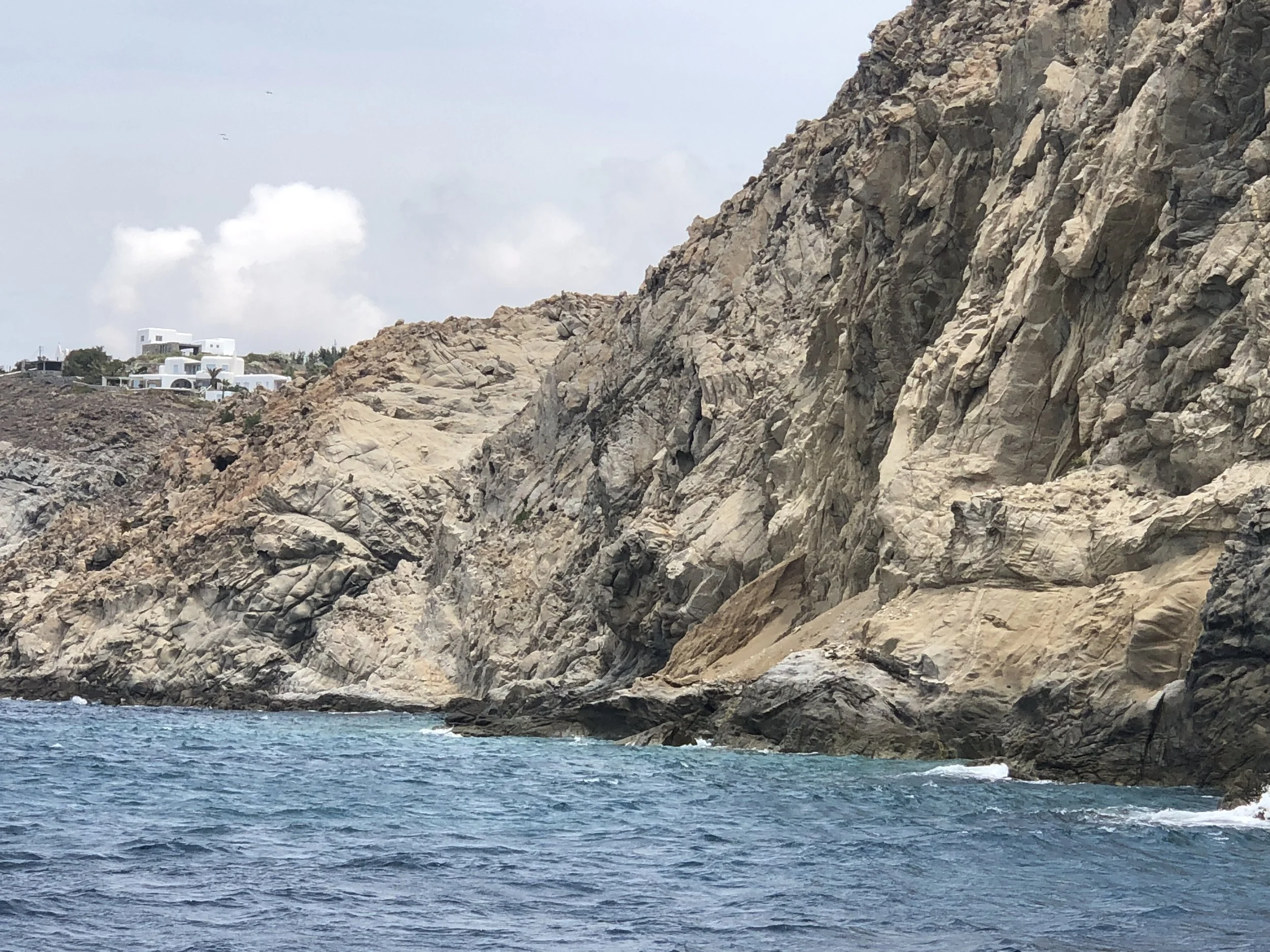 Rocky coastline with cliffs and blue sea, white buildings in the distance under a partly cloudy sky.
