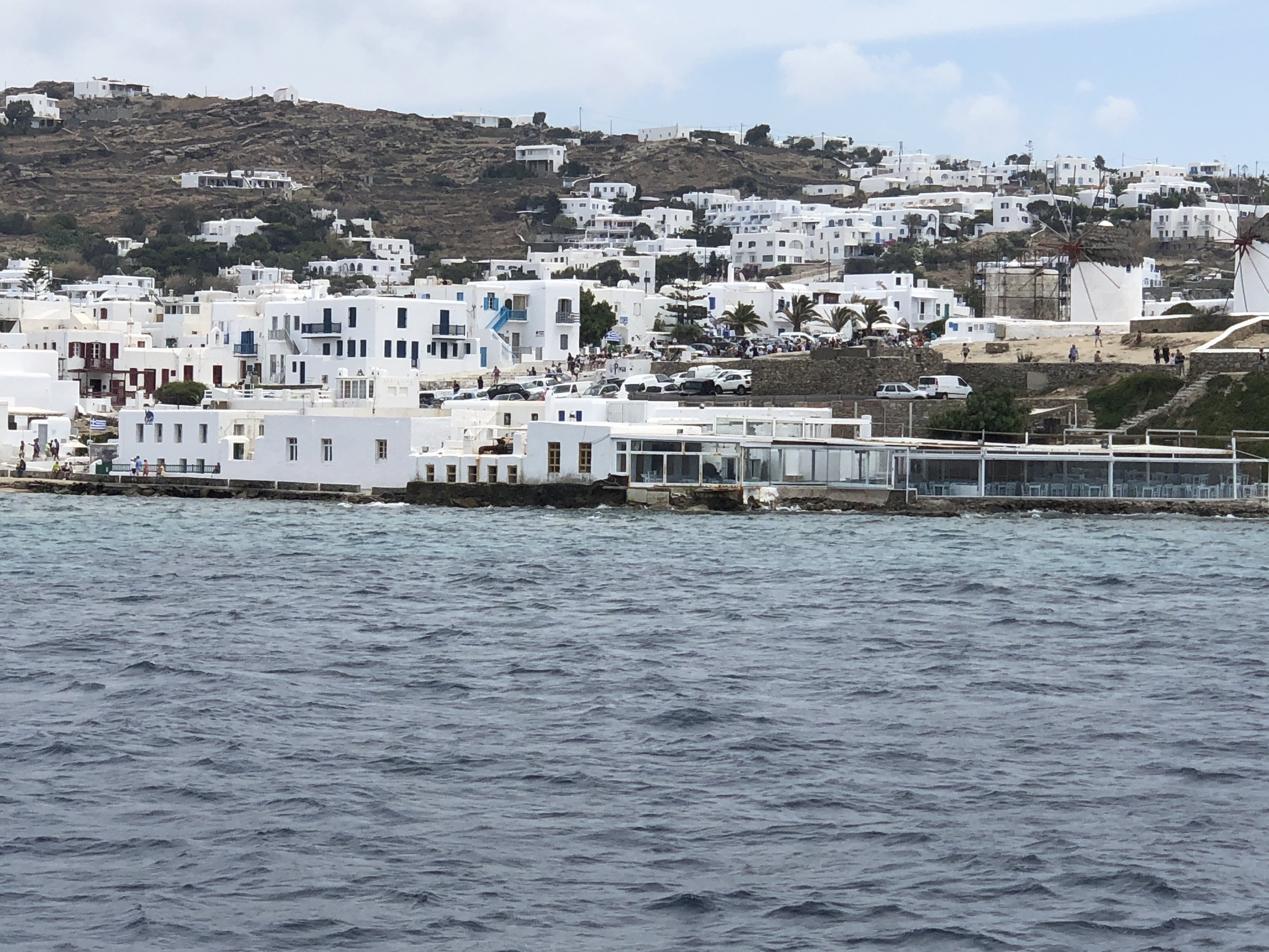 Scenic view of Mykonos town with white buildings, iconic windmills, and waterfront, Greece.