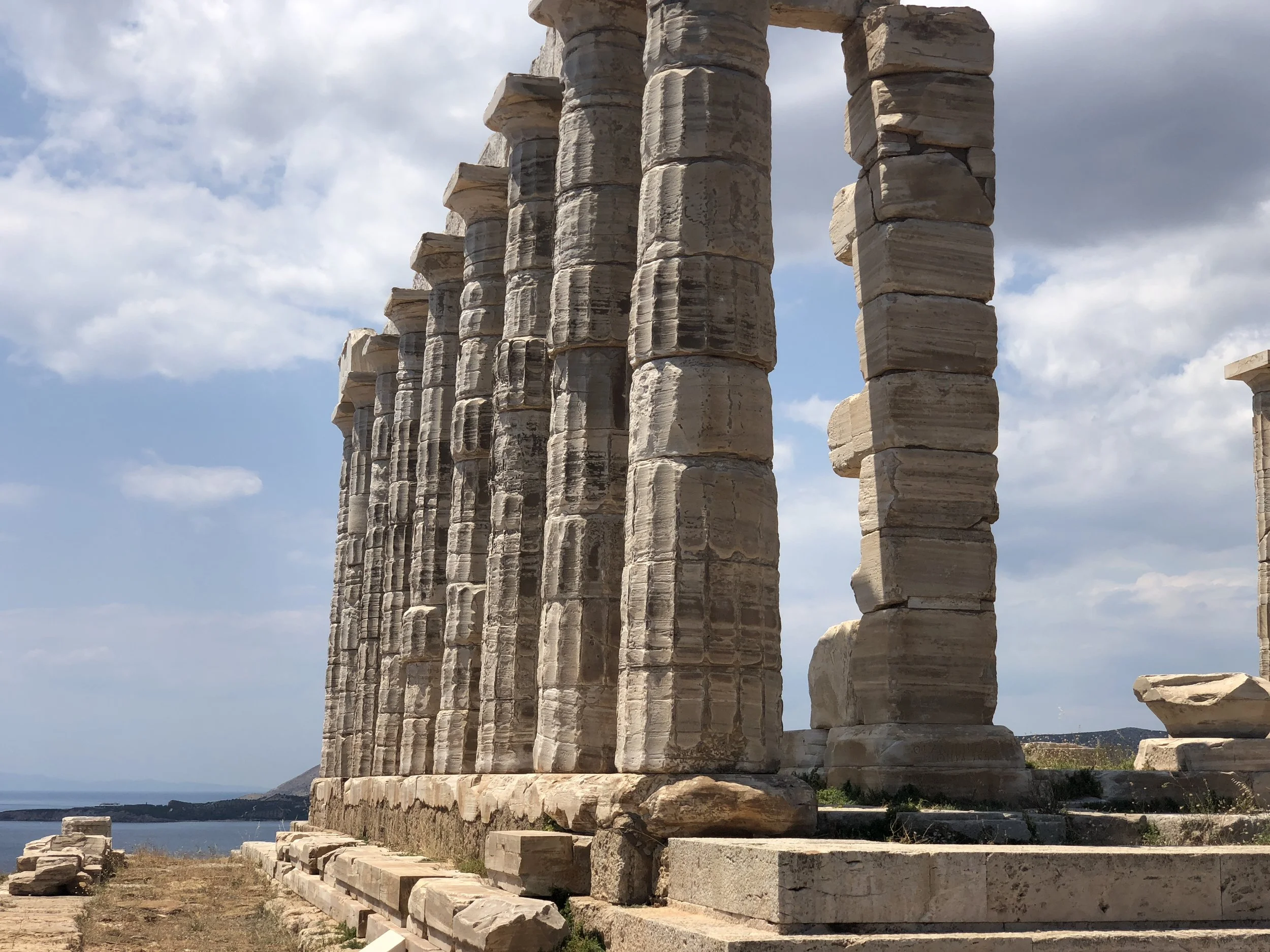 Ancient Greek temple ruins with stone columns under a cloudy sky, overlooking the sea.