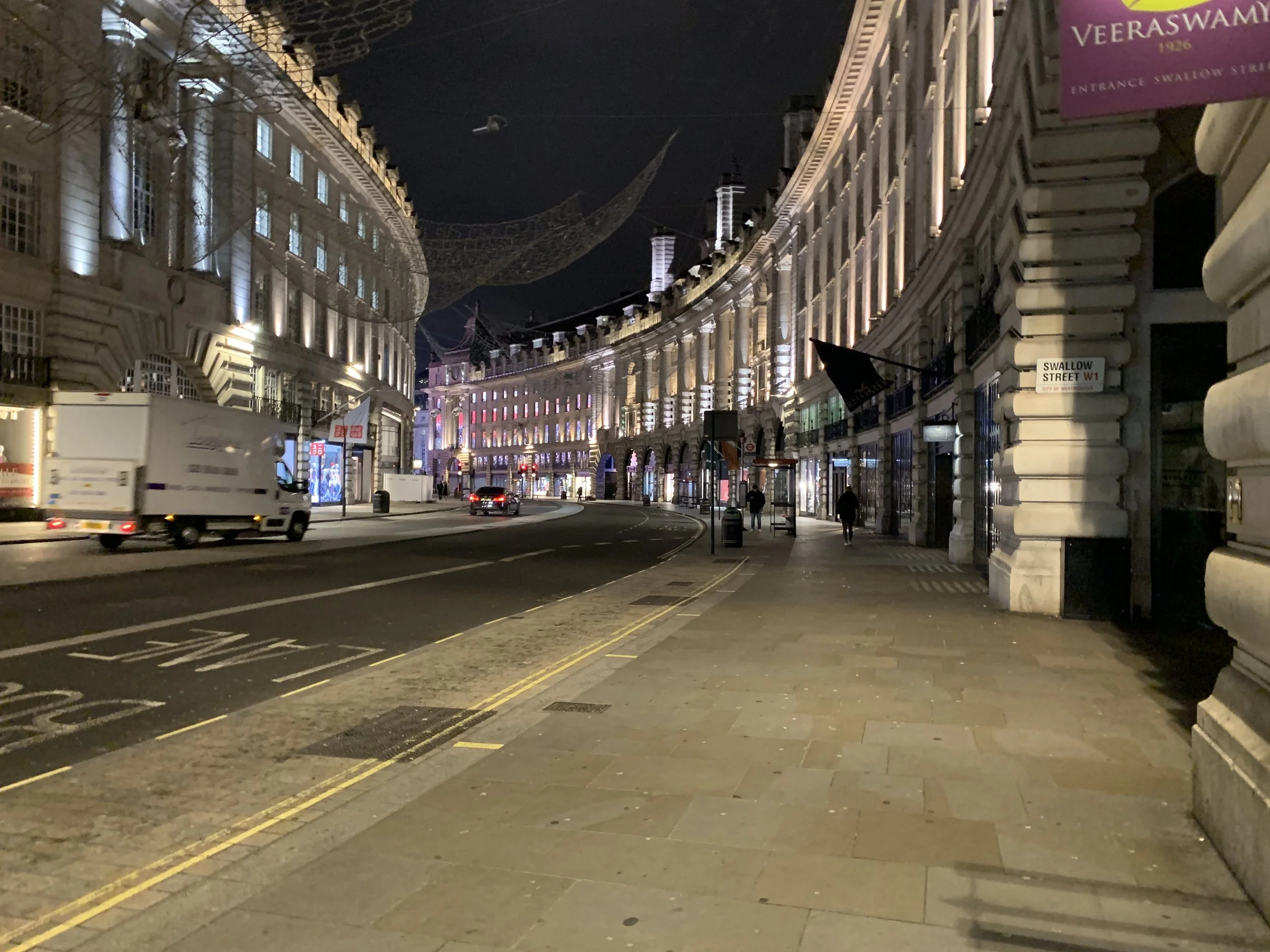 Night scene of a curved street in London with illuminated buildings, a truck and cars on the road, and a few pedestrians walking on the sidewalk.