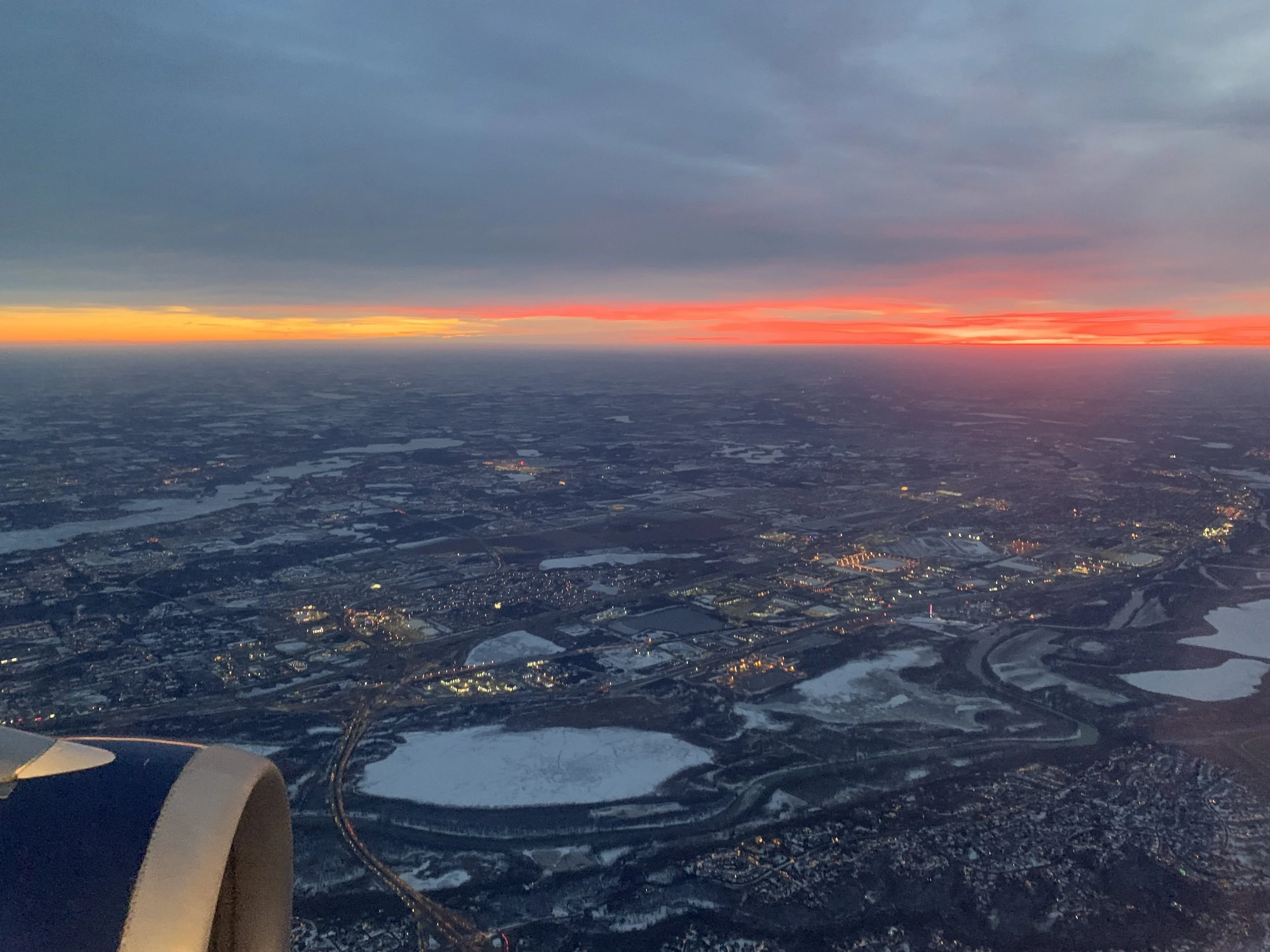 Aerial view of a cityscape with illuminated buildings at twilight, showing a mix of urban areas and natural features like rivers and lakes. The horizon displays a colorful sunset with shades of orange, red, and yellow blending into the darkening sky.