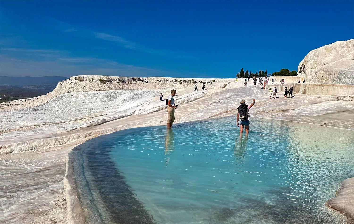 A view of an artificially constructed pool on the travertine terraces at Pamukkale, Türkiye, with two tourists wading in the pool and about 10 more tourists in the far distance.