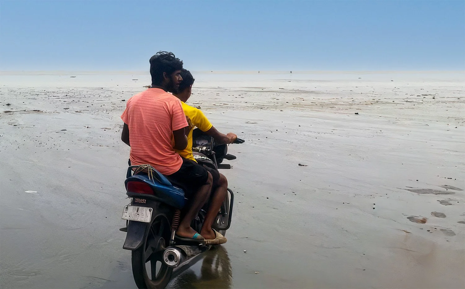 Two people riding a motorcycle on the flat, wet sand of Chandipur Beach, India, during low tide.