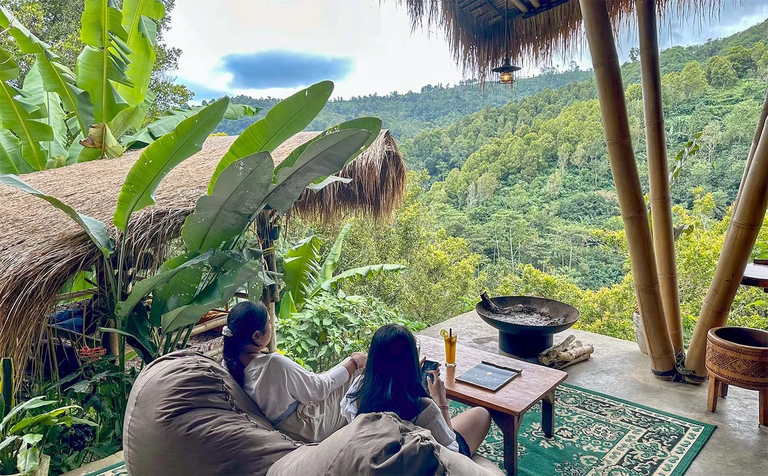 View over a forested valley in northern Bali from a restaurant with two woman sitting in the foreground on bean bags on a wooden deck.
