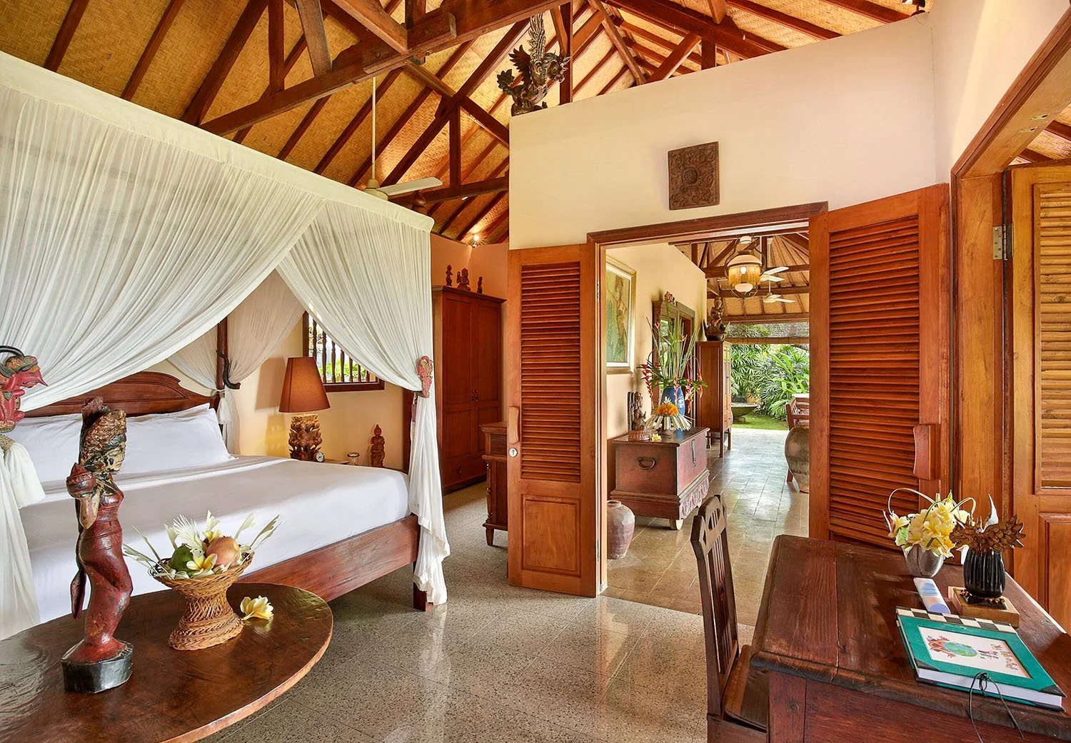 Interior of a Balinese luxury villa with high celings, timber louvered shutter doors, and a four-post canopy bed with mosquito net.
