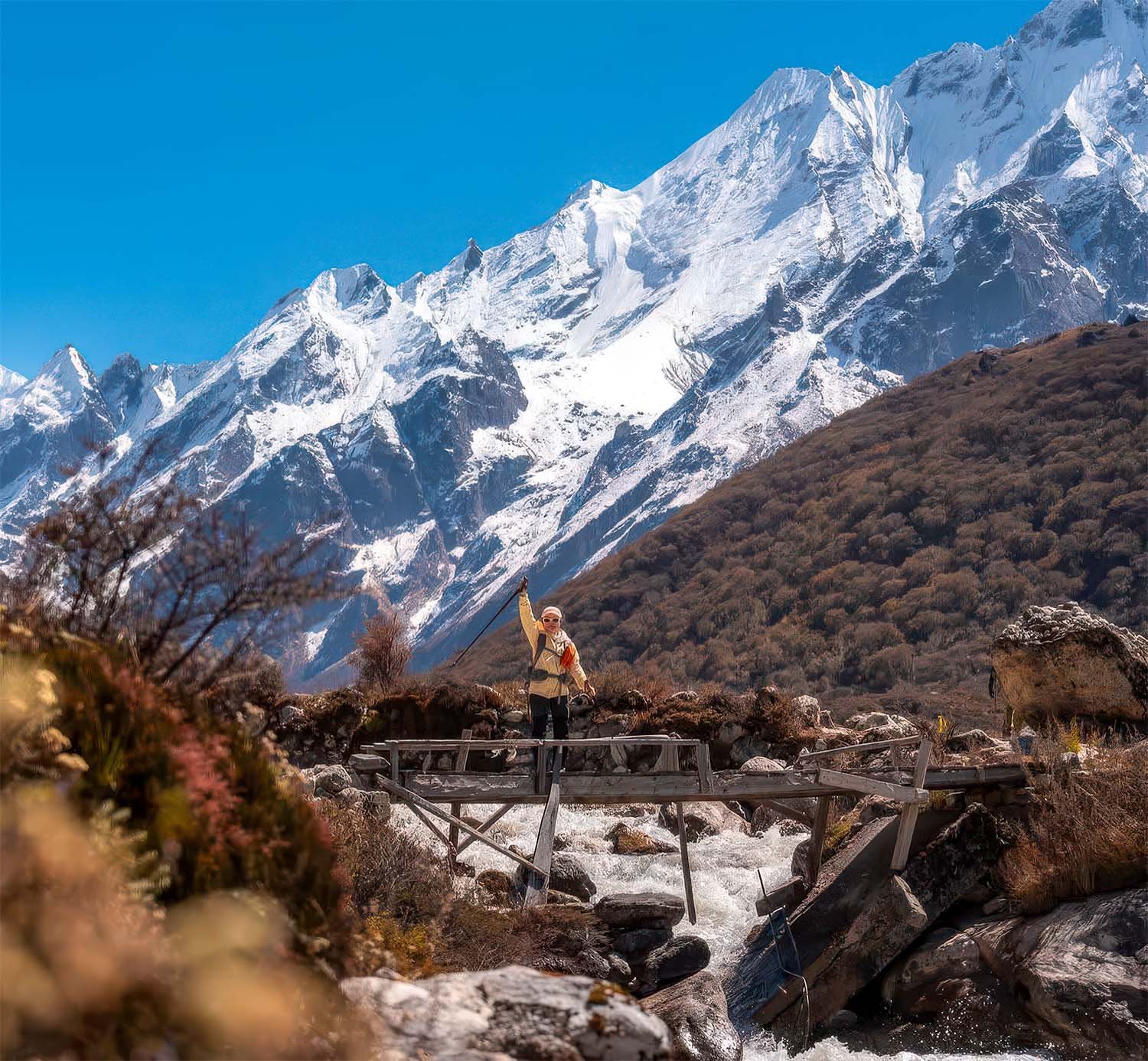 A trekker crossing a wooden bridge in Nepal with snow-covered peaks in the background.