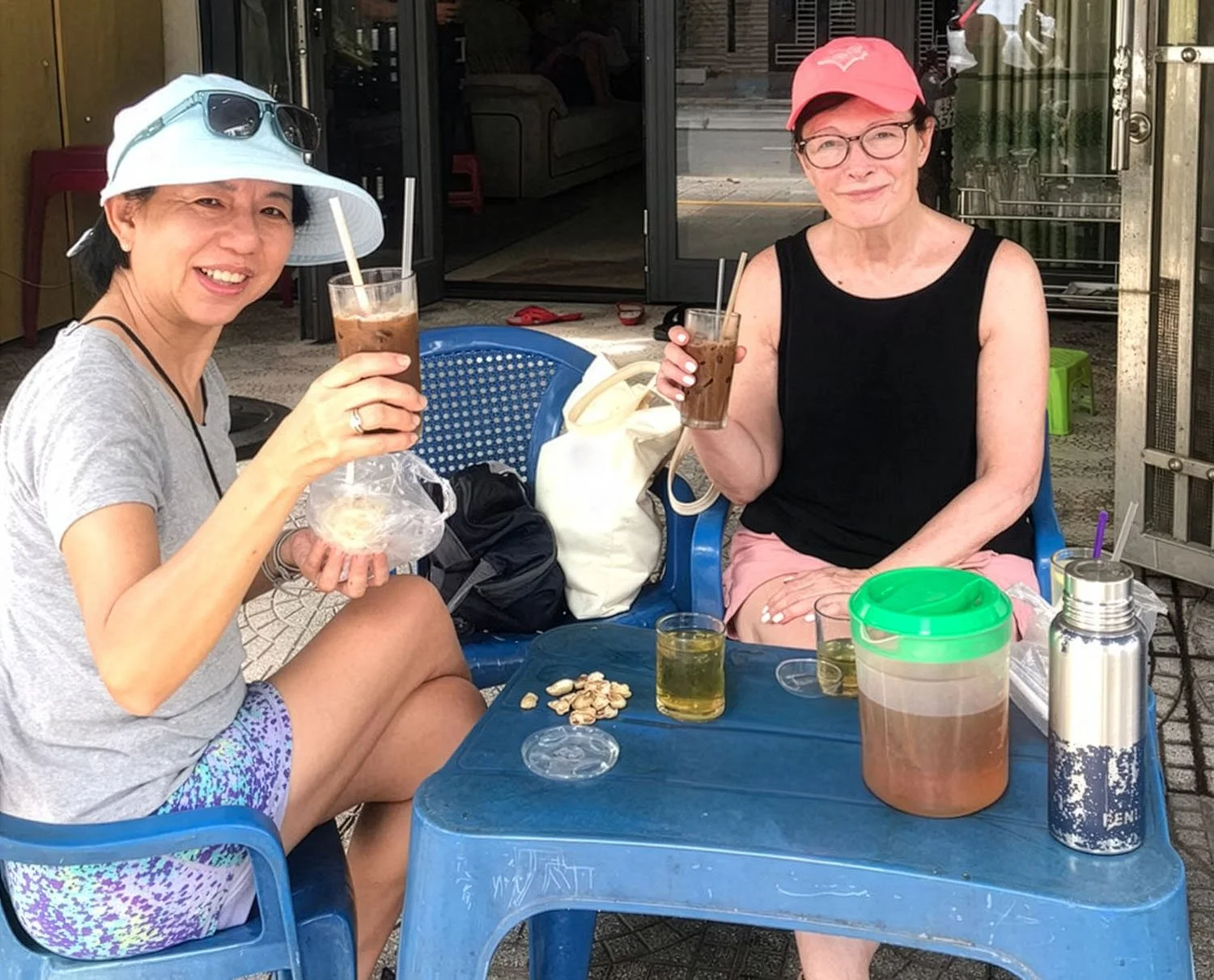 Two tourists sitting on low blue plastic chairs outside a coffee shop in Danang, Vietnam, drinking ice coffees and smiling towards the camera.