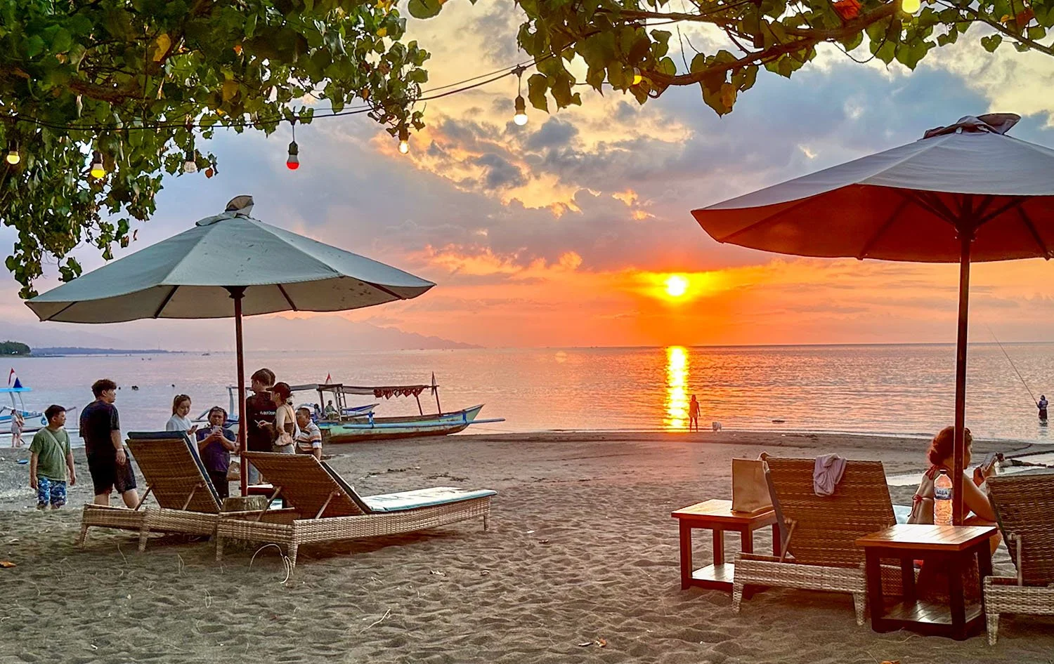 Sunset view from the Sea Breeze seafood restaurant at Lovina Beach in North Bali with two sets of sun loungers and umbrellas in the foreground and a small fishing boat in the middle distance.