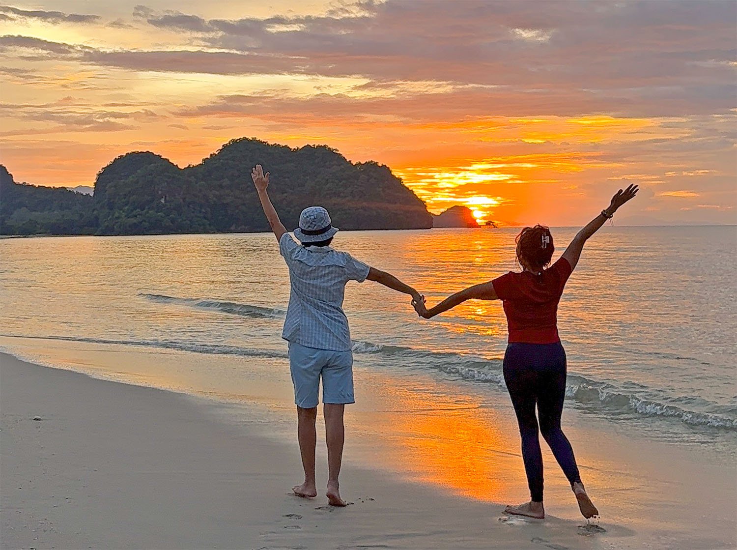 Two tourists hold hands and reach for the sky while watching a sunset from a beach on Langkawi, Island, Malaysia.