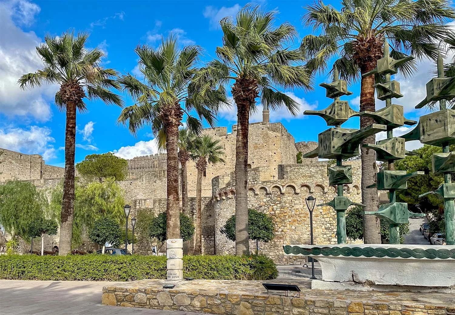 Exterior view of Çeşme Castle, Türkiye, with parkland and palm trees in the foreground;