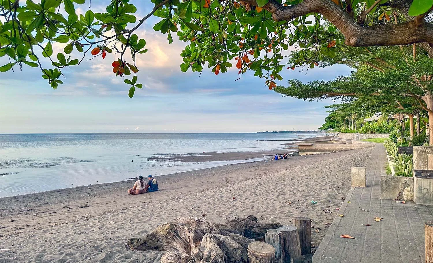 An almost empty grey sand beach at Lovina Beach in North Bali with a concrete pathway along the top of the beach.