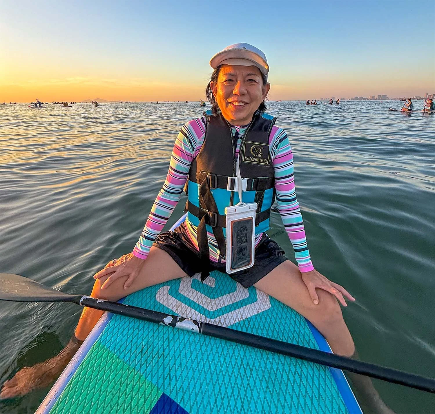 Female Asian tourist sitting on a paddleboard in the ocean at Danang, Vietnam, facing the camera with a sunset behind and other paddlers in the distance.