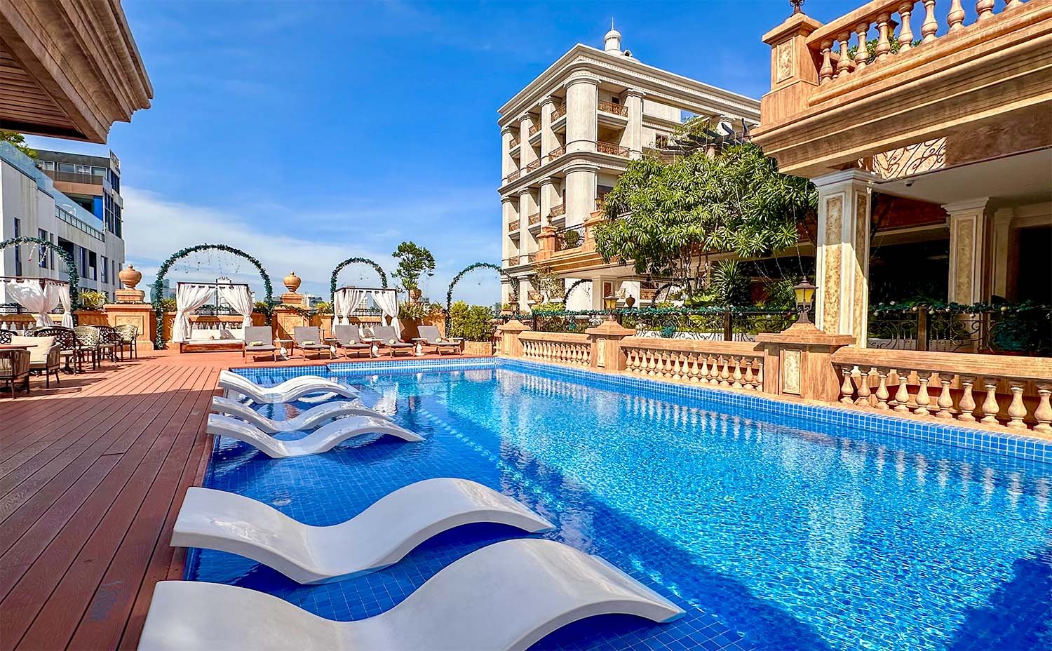Wide view of a large swimming pool on the rooftop of the Anik Palace Hotel in Phnom Penh, Cambodia, with white recliner chairs on the edge of the pool and a neo-classical design building in the background.