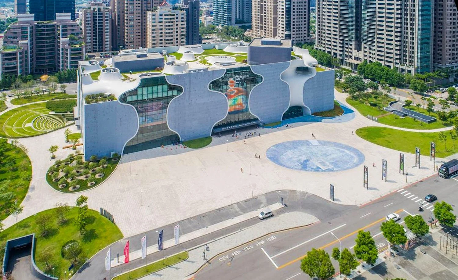Semi-aerial view (from a nearby high rise building) of the National Theatre in Taichung, Taiwan, and surrounding parkland with office and residential high rise buildings in the background.