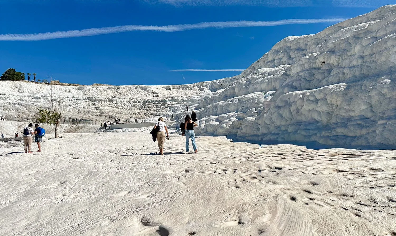 View up the travertine terraces in Pamukkale, Türkiye, with four tourists in the middle distance and about 12 tourists in the far distance.