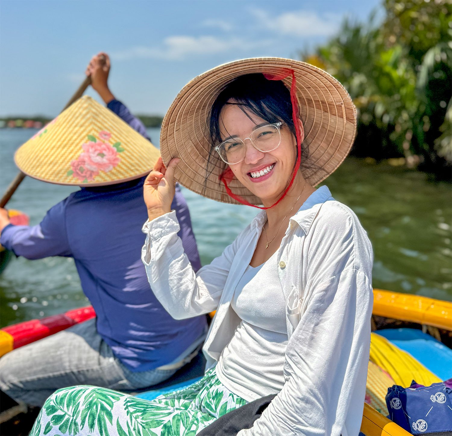 A middle-aged Vietnamese woman wearing a traditional nón lá hat smiles towards the camera whilst sitting in a round bamboo boat on a river behind the  'driver' who is steering it with a wooden paddle.