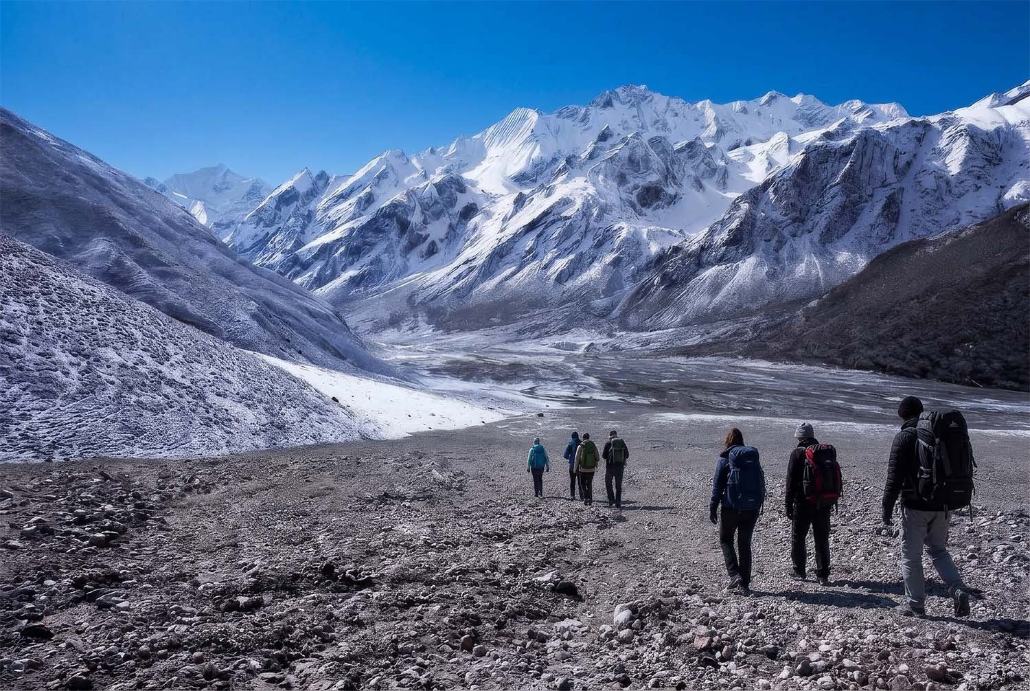 Seven trekkers cross a rocky plateau in the upper reaches of the Himalayan Mountains of Nepal walking towards a wide valley surrounded by snow-covered mountains.
