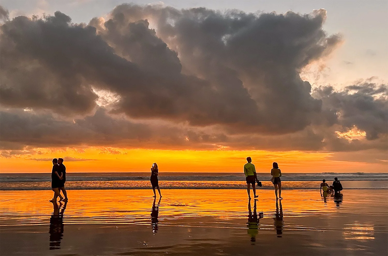 Tourists walking on the beach in Seminyak, Bali, with several taking photos of the sunset.