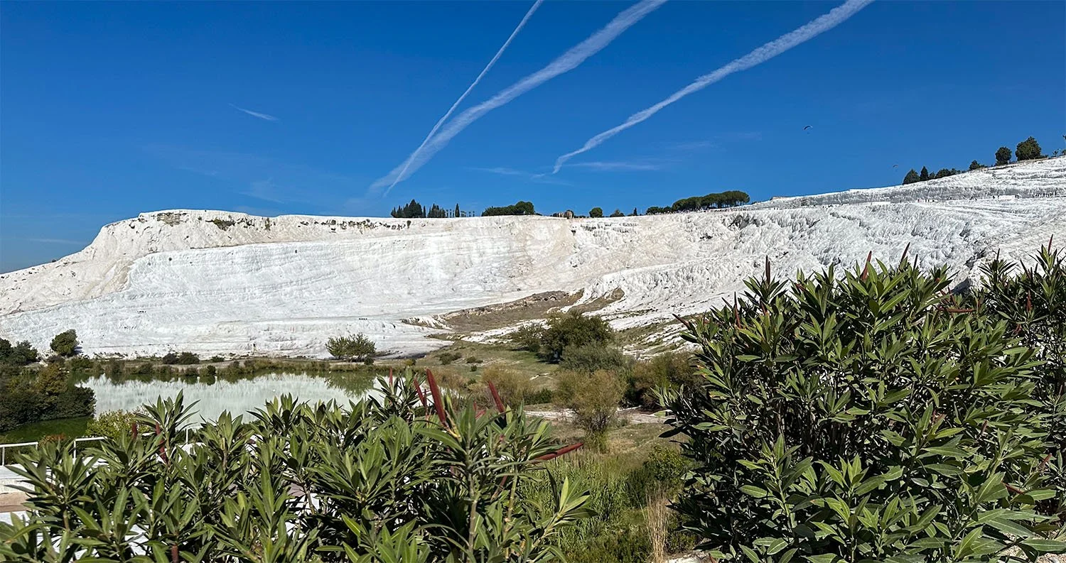 A distant view of the Pamukkale travertine terraces in Türkiye against a blue sky with aircraft contrails.