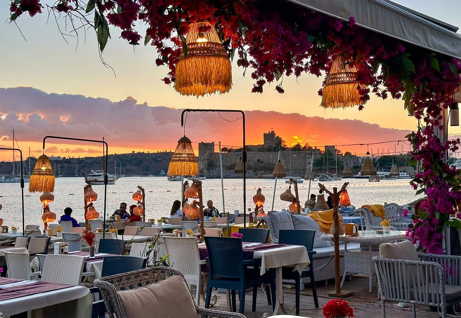 View of a waterfront restaurant in Bodrum, Turkey, with silhouette of Bodrum Castle against a setting sun in the background.
