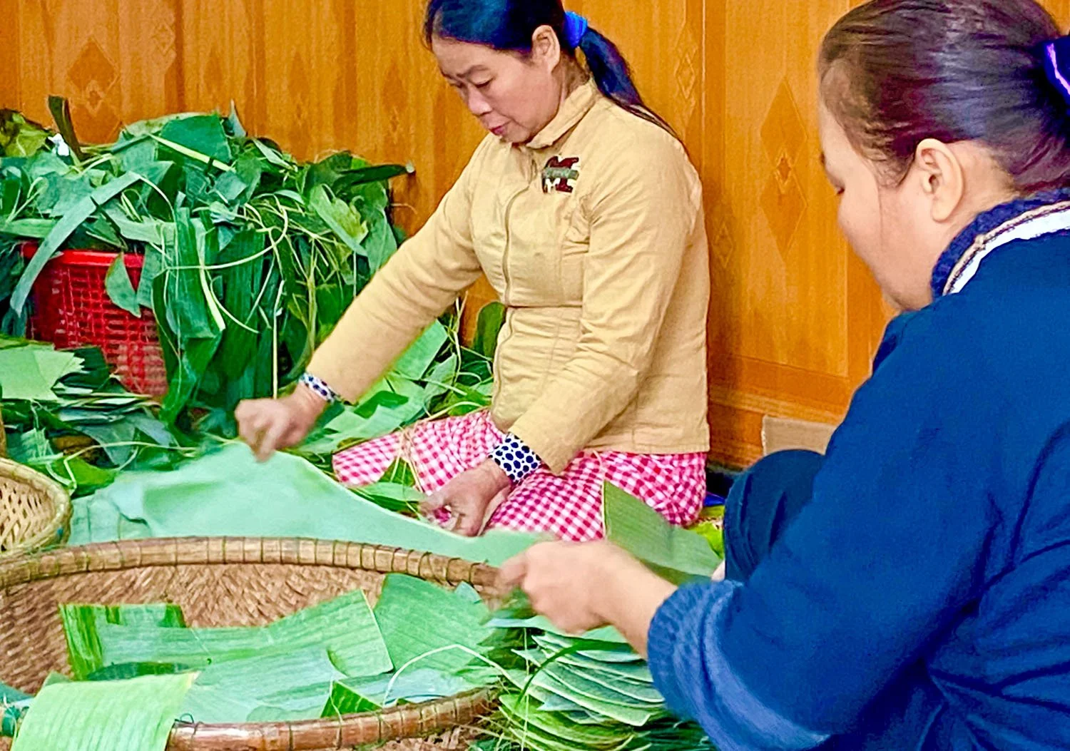 Two Vietnamese women sitting on the floor cut banana leaves into shapes for wrapping Tet cakes.