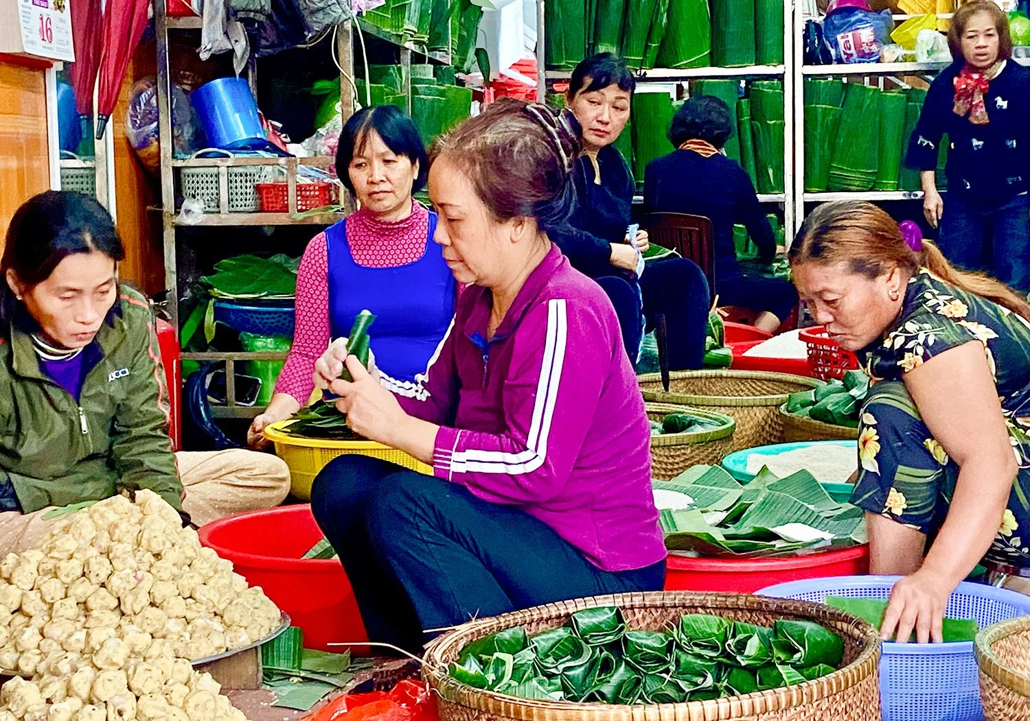 Seven middle-aged women sitting on the floor of a large kitchen making Tet cakes.