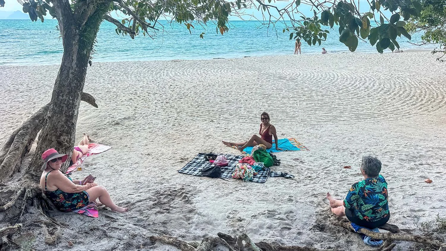 Three older women relaxing on beach towels on an empty white sand beach on Langkawi Island, Malaysia.