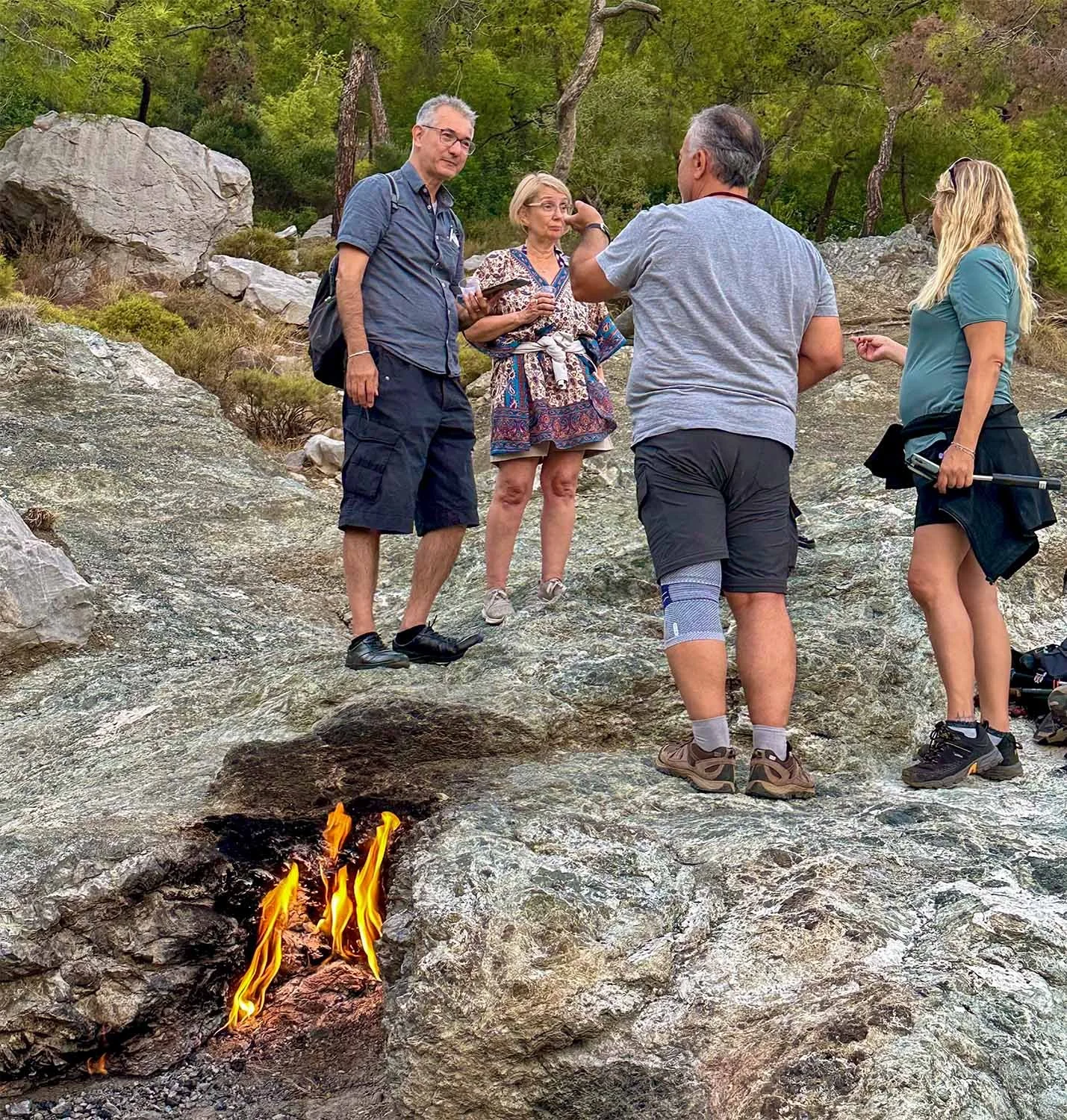 Four tourists chatting on Mount Chimaera, Türkiye, standing next to one of the eternal flames burning from fissures in the rock near their feet.
