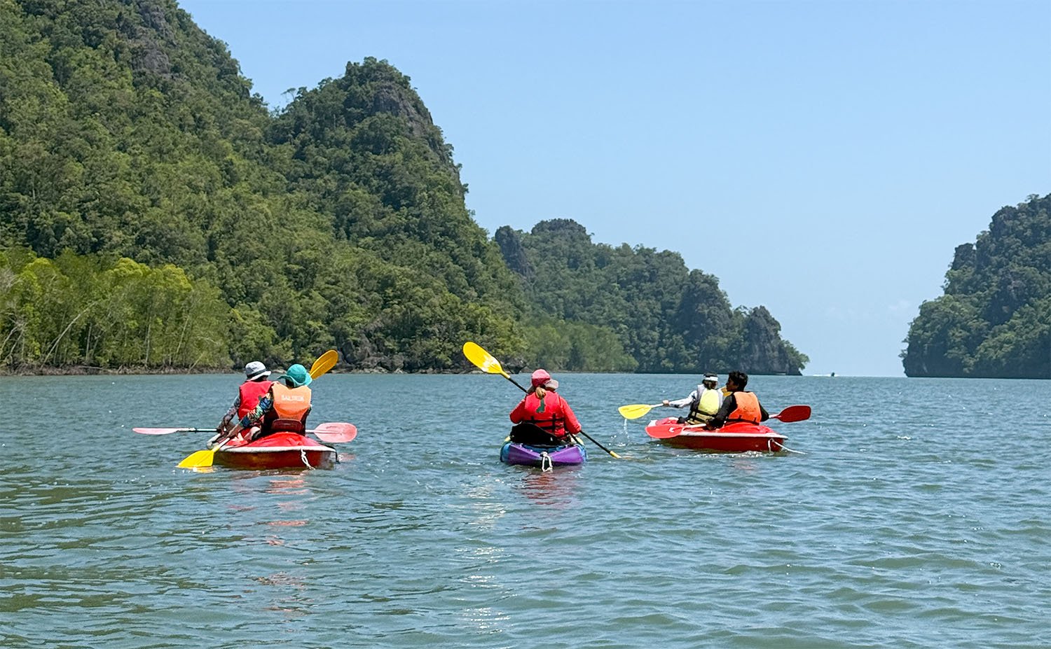 Five tourists in lifejackets paddle three kayaks on a smooth ocean in the Kilim Geopark on Langkawi Island, Malaysia.