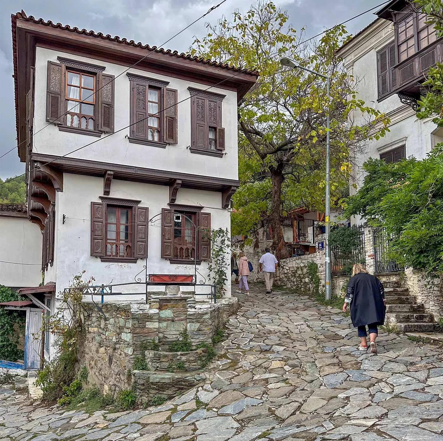 Three tourists walk up a narrow cobblestone street between traditional house with intricately carved wooden shutters in Şirince, Türkiye.