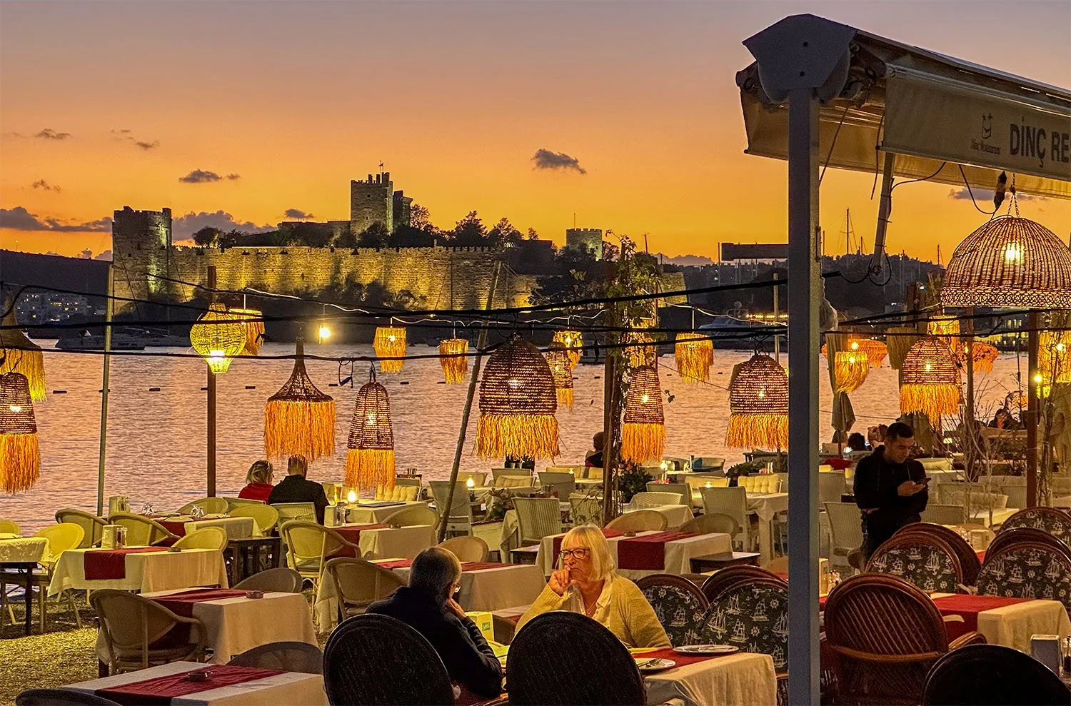 View across a waterfront restaurant at Bodrum, Türkiye, with Bodrum Castle in the background behind which the sun has just set.